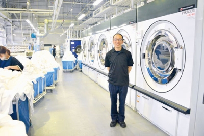 写真：最新の大型洗濯機や乾燥機が並ぶローカルベース登別工場と田島工場長